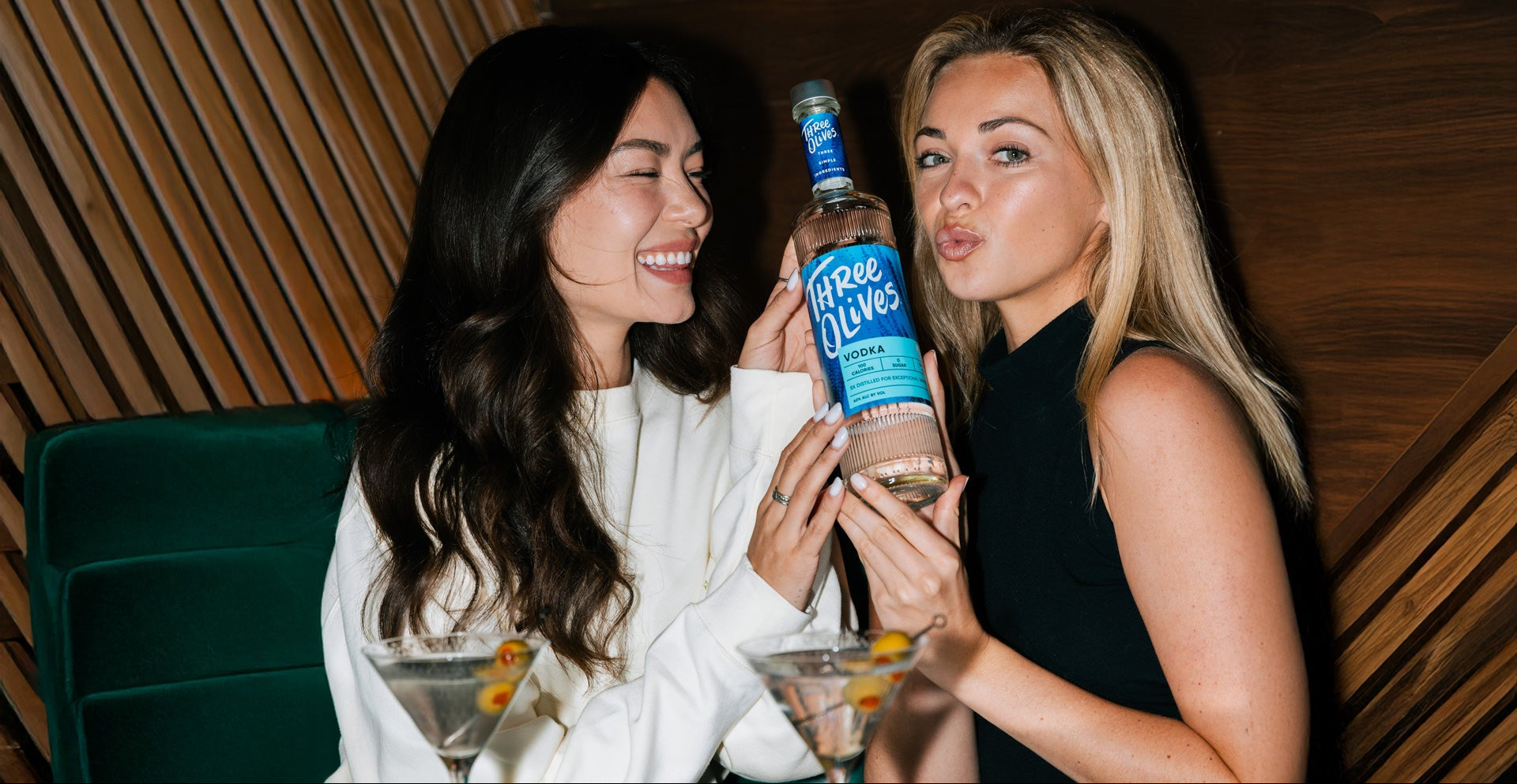 Two women holding a bottle of Three Olives Vodka original flavor at a social gathering.